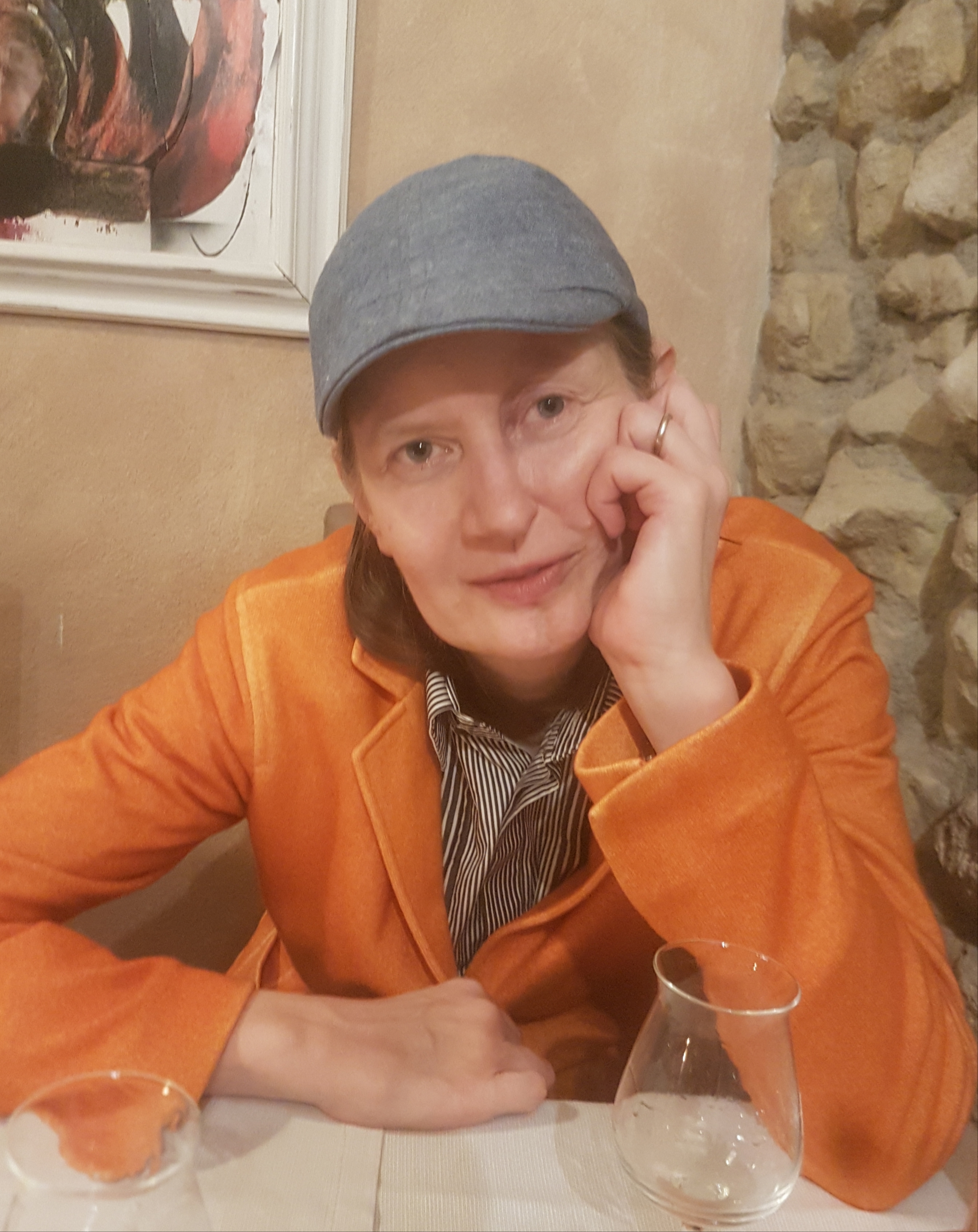 Autrice Elena Nikulina, con giacca arancione e berretto grigio chiaro, seduta a un tavolo in posa riflessiva con la.testa appoggiata alla mano.